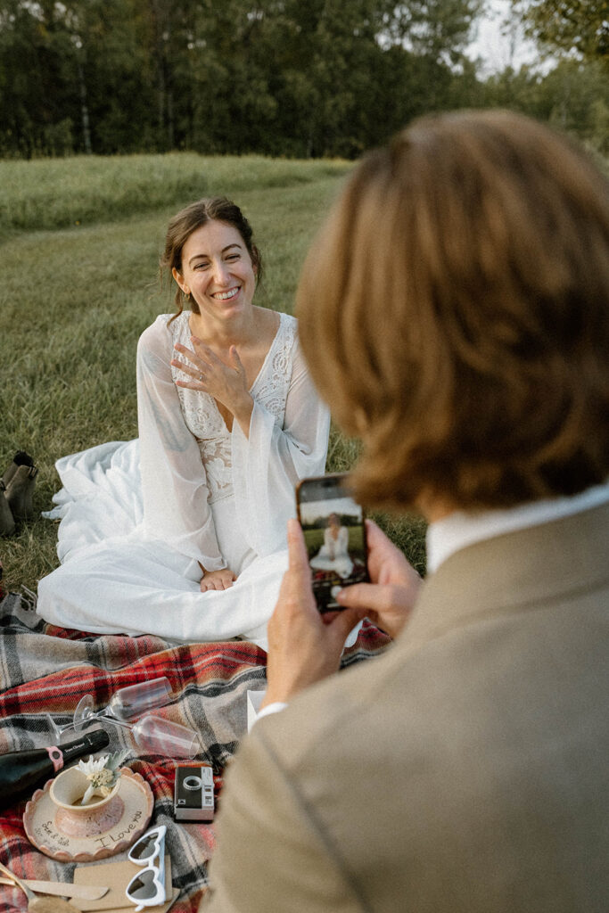 Bride sitting on a picnic blanket showing off her wedding ring while the groom takes a photo during a relaxed Vermont elopement celebration outdoors.