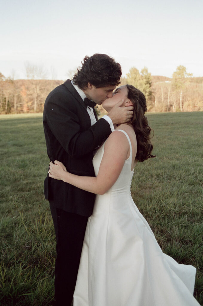 Bride and groom in formal attire share a kiss in a grassy Vermont field at golden hour, rolling hills and autumn trees in the background.