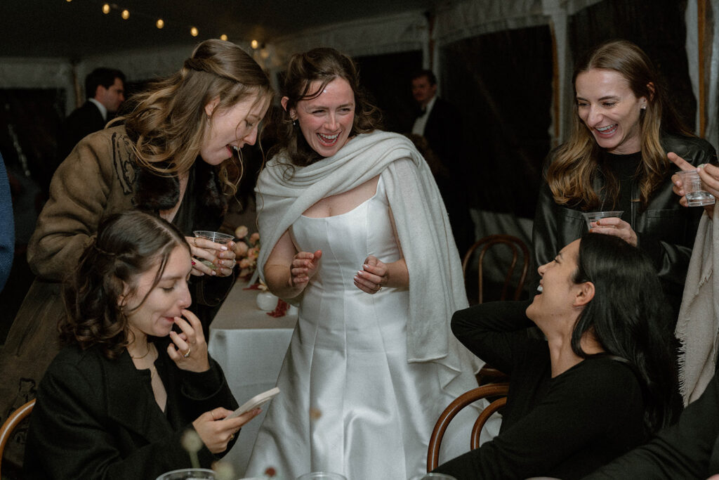 Bride wrapped in a shawl laughs with friends gathered around a table during the evening reception.