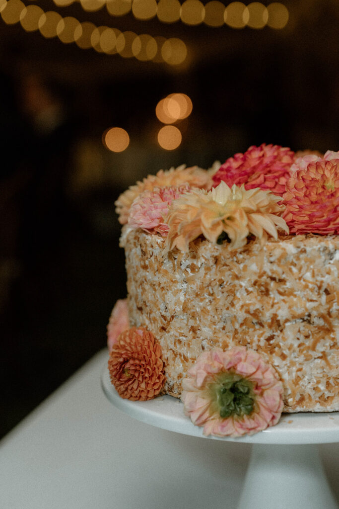 Small coconut wedding cake decorated with fresh dahlias sits on a white cake stand with string lights glowing in the background.