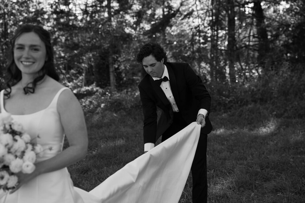 Groom in a black tuxedo lifts the bride’s long train as she walks ahead through a wooded clearing.