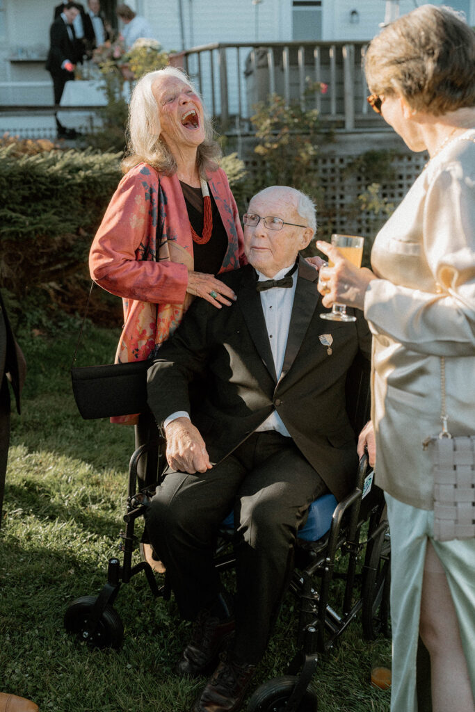 Elderly couple laughing together during backyard Vermont wedding cocktail hour in Greensboro with family gathered around.