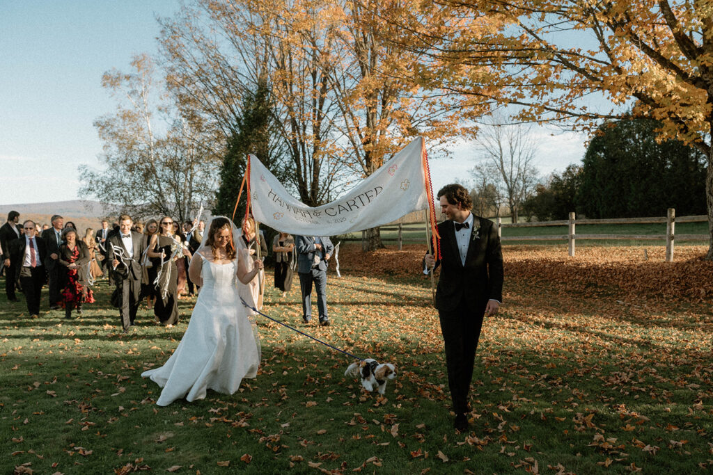 Bride and groom lead wedding guests across a field holding an embroidered “Hannah & Carter” banner while walking their dog.