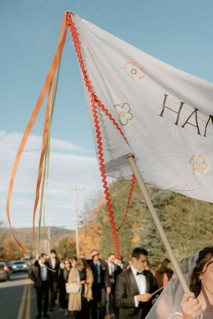 Close-up of an embroidered wedding banner with ribbon streamers reading “Hannah & Carter,” carried during a wedding procession.
