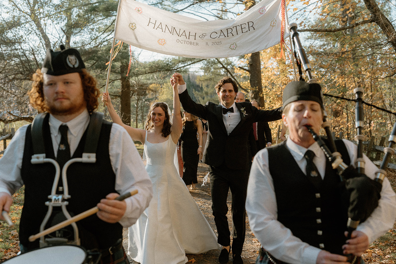 Newlyweds celebrating beneath a banner while a bagpipe band leads the joyful wedding parade through the Vermont countryside.