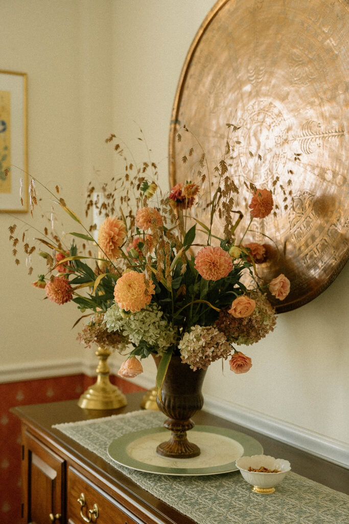 Fall floral arrangement with peach dahlias, grasses, and hydrangea in a brass vase on a wooden sideboard beneath a large antique copper tray.