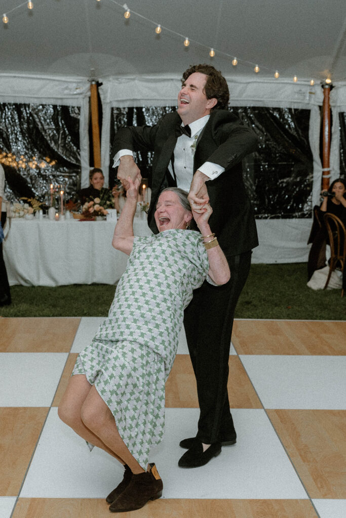 Groom spins an older woman joyfully across the dance floor inside a tented wedding reception.