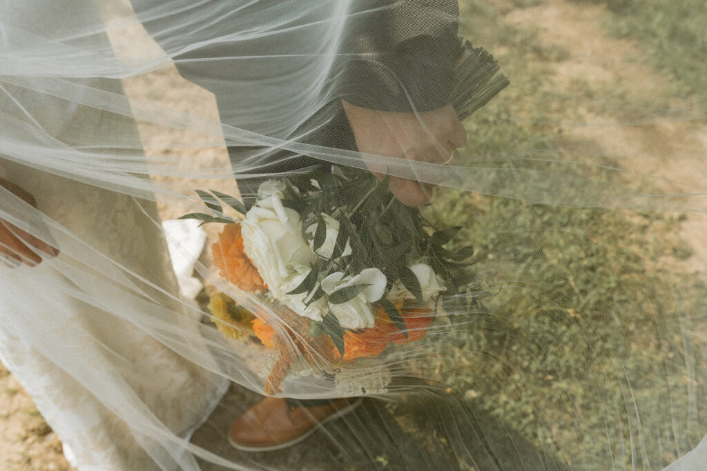 Bride and groom standing close together while holding the bride’s bouquet beneath a flowing veil in warm evening light.