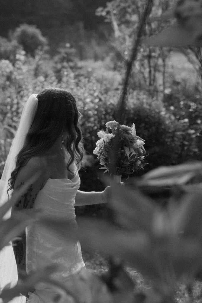 Black and white photo of the bride walking through a lush Vermont garden holding her bouquet with sunlight filtering through the trees.