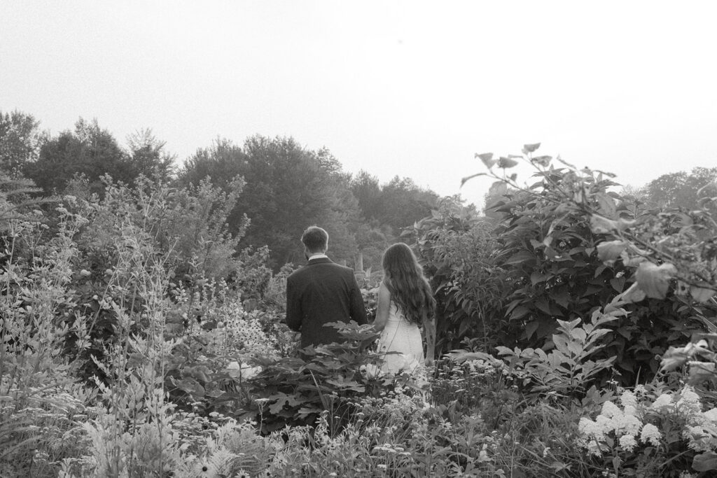 Bride and groom walking away together down a garden path through tall summer flowers and greenery after their ceremony.