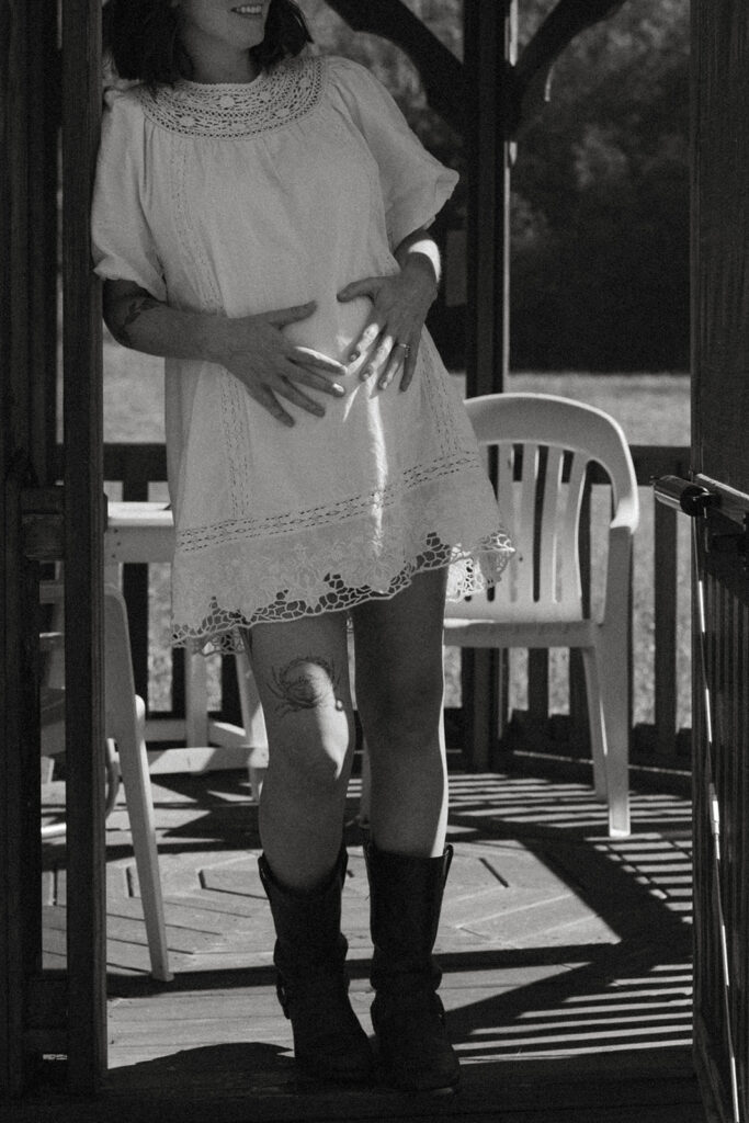 Black and white photo of a pregnant woman gently holding her belly in a lace dress inside a gazebo