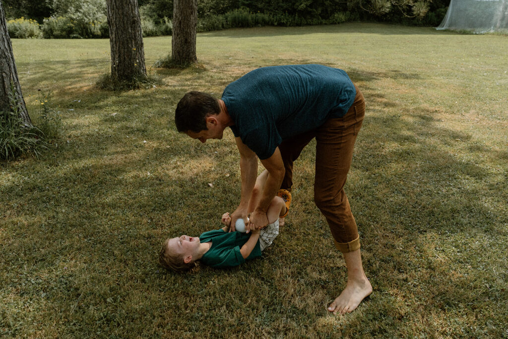Dad playfully tickling his young son on the grass during relaxed backyard family photos at home in Vermont.