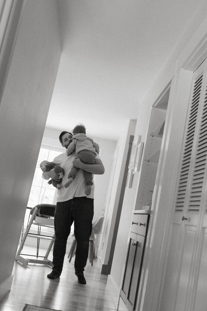 Dad walks through a bright hallway holding baby and toddler during a documentary-style family photo moment
