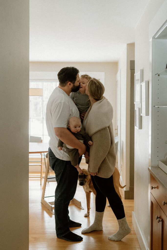 Parents kiss their toddler while holding baby in a warm, light-filled kitchen during a lifestyle family session