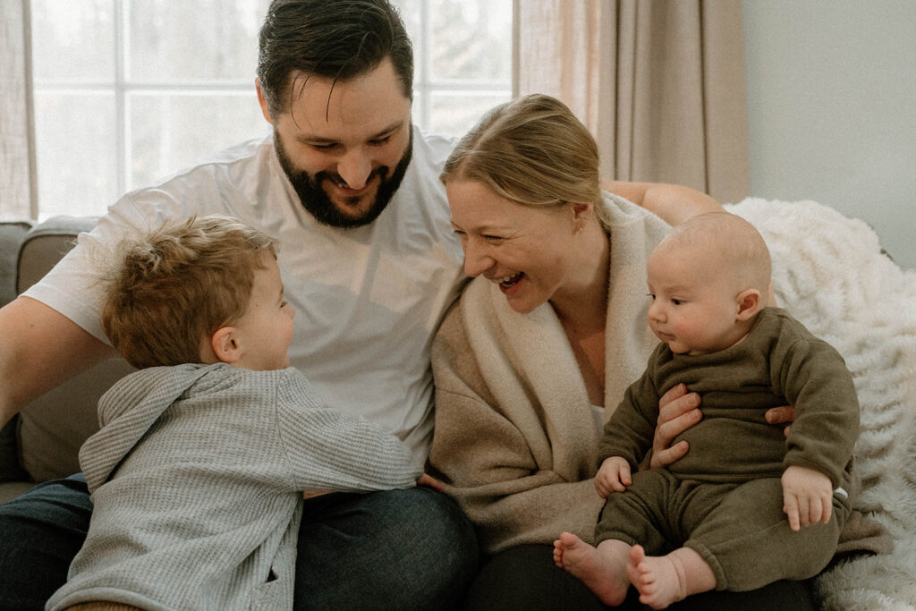 Parents sit on the couch laughing with their two young children during a relaxed lifestyle family photo session at home