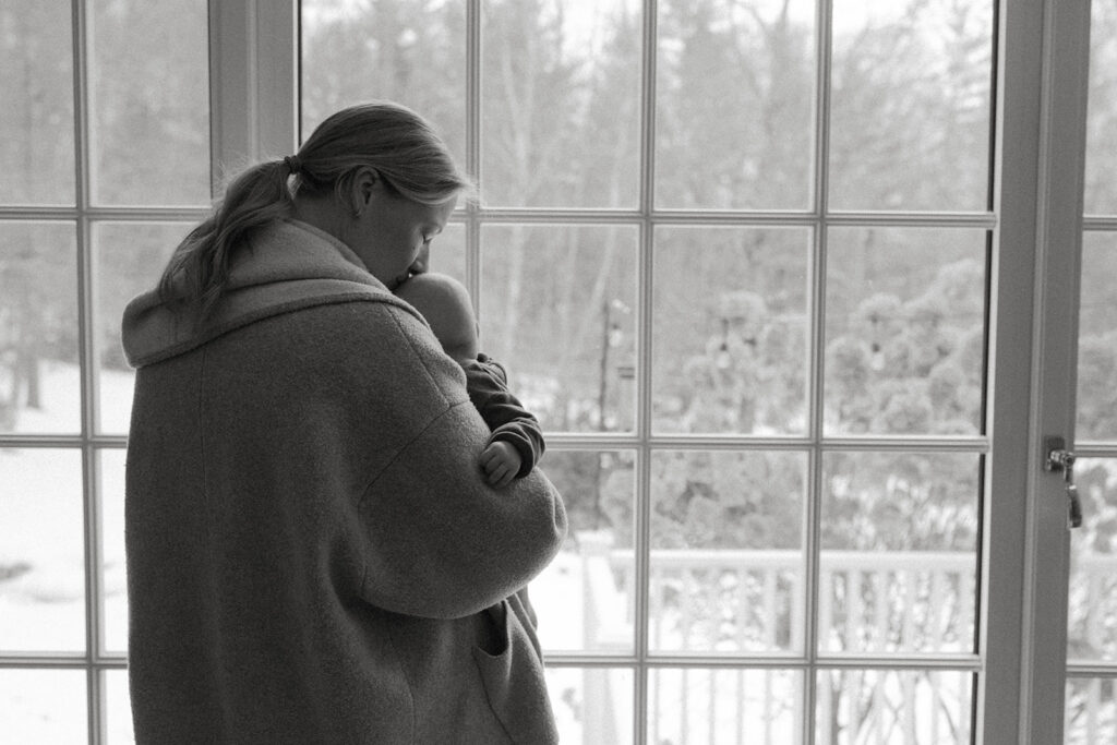 Mom holds baby by a large window with snowy Vermont backyard view during a quiet winter family photo moment