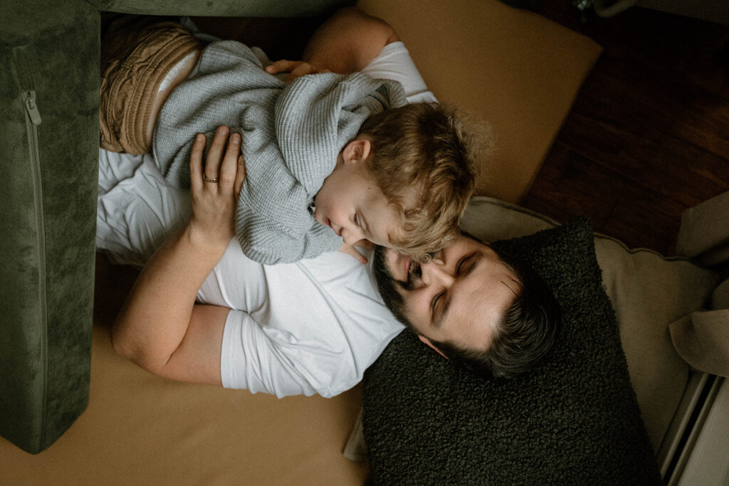 Dad lies on the couch with toddler curled into him during a candid in-home family photo session
