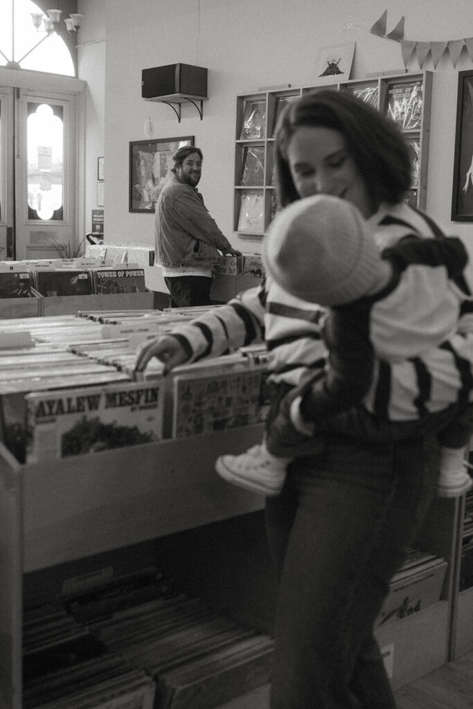 Mom and dad browse records while baby watches from mom’s arms in a cozy record shop family photo session
