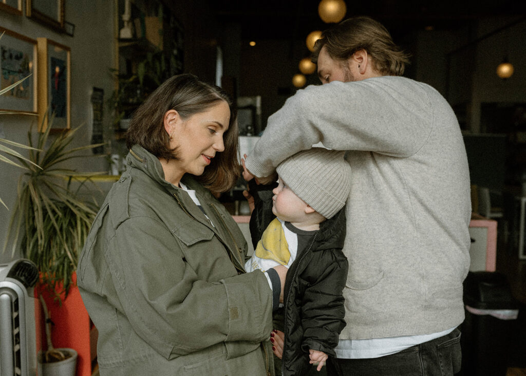 Mom zips baby into a jacket inside a warm, plant-filled coffee shop during a candid family photo session